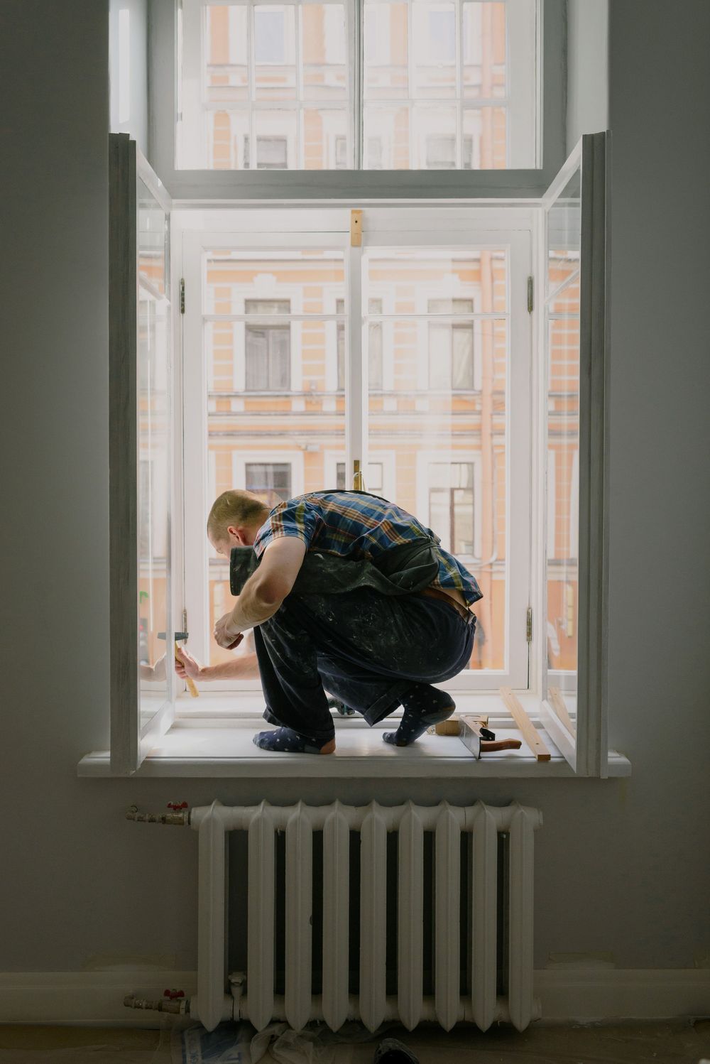 Vakman zit gehurkt in een hoog open raam en werkt aan het kozijn, met een radiator onder de vensterbank